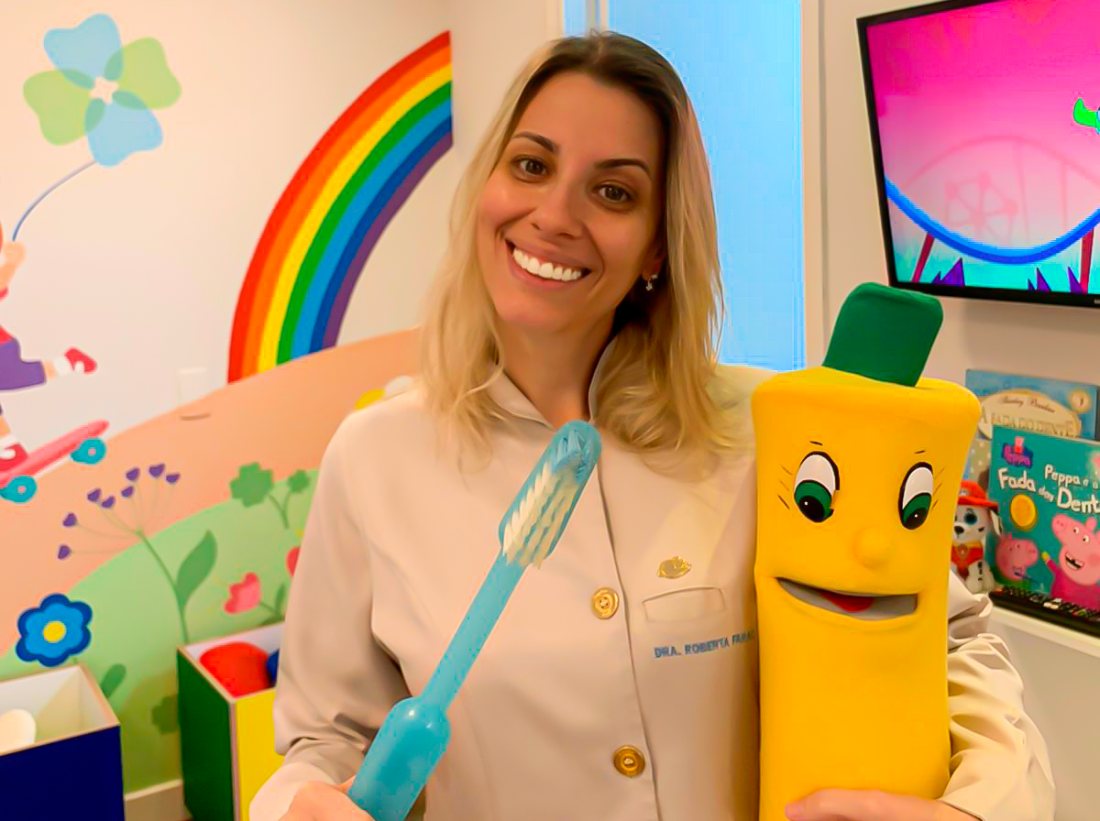 Dra. Roberta Farac sorrindo, ao lado do mascote escova de dentes, na sala infantil